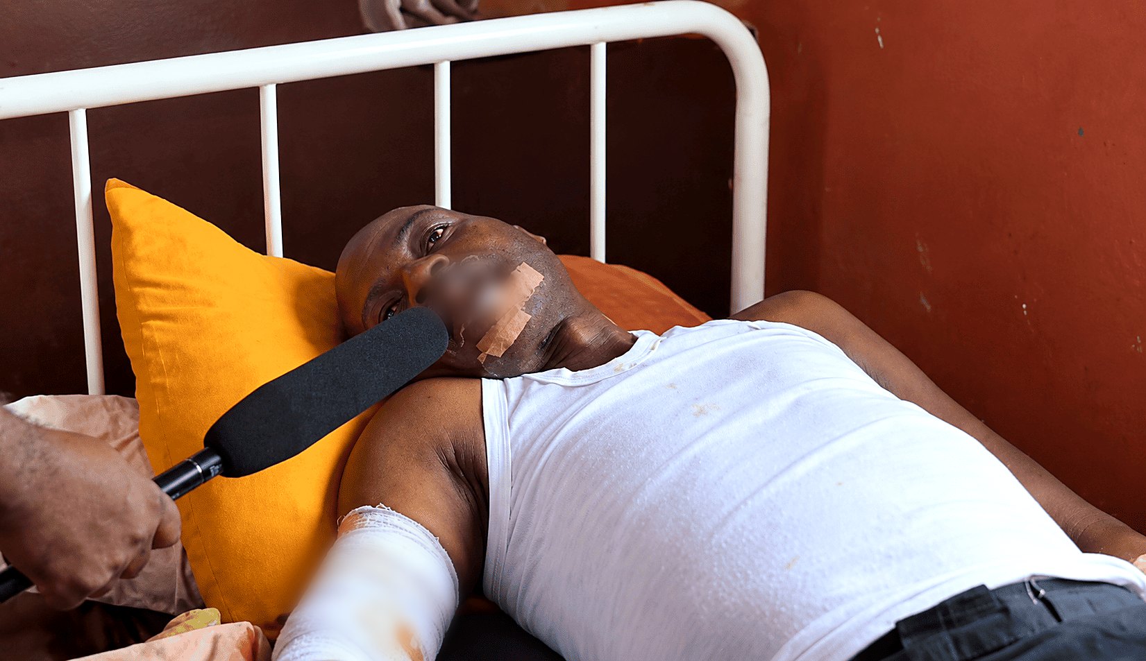 Anthony Bulus lying in a hospital bed with bandaged wounds, speaking to a reporter
